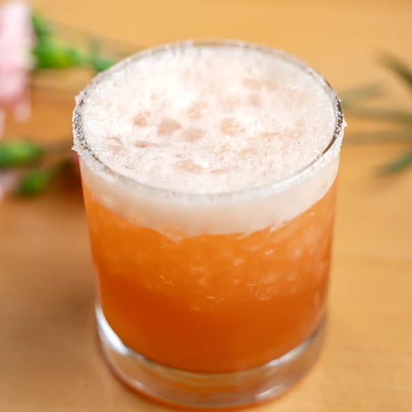 A vibrant orange cocktail with a foamy top and a sugar-rimmed glass on a wooden table.