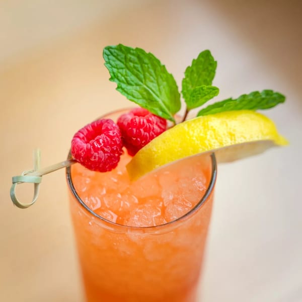A vibrant orange cocktail with crushed ice, garnished with mint, lemon, and raspberries.