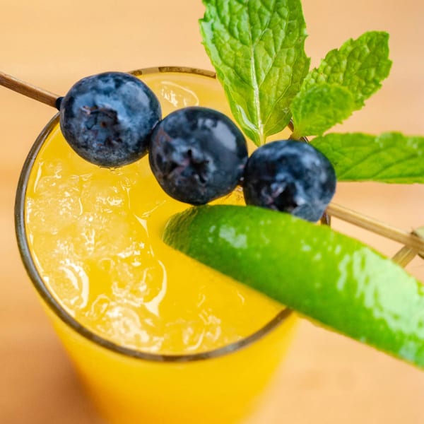 Close-up of a vibrant yellow drink with ice, garnished with blueberries, mint, and a lime wedge.