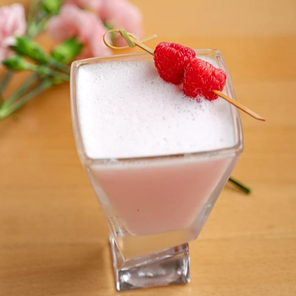 A pink, foamy cocktail garnished with two raspberries on a skewer, with pink flowers in the background.