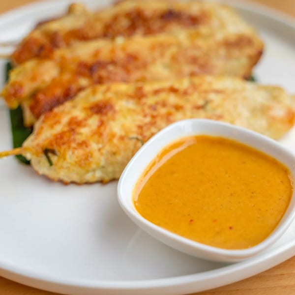 Three golden-brown chicken satay skewers with a side of orange peanut dipping sauce on a white plate.